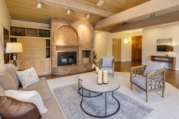 A cozy living room with wooden beams on the ceiling, a brick fireplace, and a mix of modern and rustic furniture. There is a beige sofa with accent pillows, two armchairs with a lattice design, and a round coffee table holding candles. The walls are painted in a light neutral tone and the flooring is wooden. A large rug adds warmth to the space. Shelving units and a small table with a lamp provide additional decor.