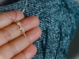 A hand holding a silver necklace with a crucifix pendant. The background is a textured fabric with shades of blue and black.