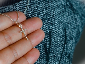A hand holding a silver necklace with a crucifix pendant. The background is a textured fabric with shades of blue and black.