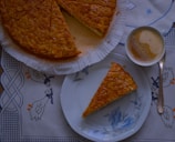 A piece of cake with an almond topping is placed on a floral-patterned plate alongside a cup of frothy coffee on a decorative tablecloth featuring blue and orange ducks. The rest of the cake is partially visible, still on its parchment-lined base.