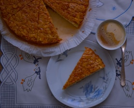 A piece of cake with an almond topping is placed on a floral-patterned plate alongside a cup of frothy coffee on a decorative tablecloth featuring blue and orange ducks. The rest of the cake is partially visible, still on its parchment-lined base.