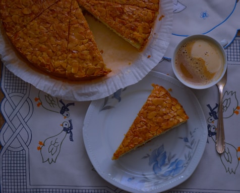 A piece of cake with an almond topping is placed on a floral-patterned plate alongside a cup of frothy coffee on a decorative tablecloth featuring blue and orange ducks. The rest of the cake is partially visible, still on its parchment-lined base.
