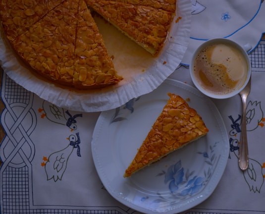 A piece of cake with an almond topping is placed on a floral-patterned plate alongside a cup of frothy coffee on a decorative tablecloth featuring blue and orange ducks. The rest of the cake is partially visible, still on its parchment-lined base.