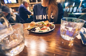 A plate of food is on a wooden table, garnished with colorful vegetables, cheese, and bread. A person wearing a dark shirt with white lettering is seated at the table. There are glasses of water and a knife and fork nearby. The background is a dimly lit restaurant with other patrons.