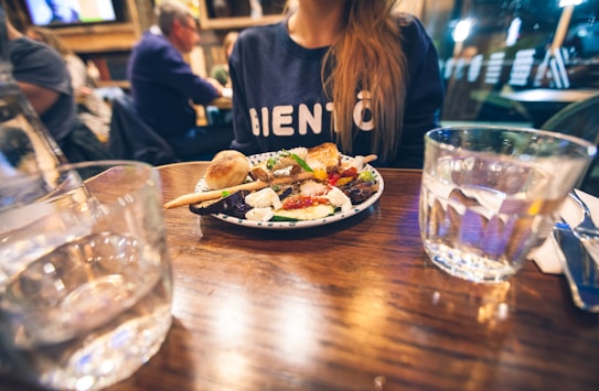 A plate of food is on a wooden table, garnished with colorful vegetables, cheese, and bread. A person wearing a dark shirt with white lettering is seated at the table. There are glasses of water and a knife and fork nearby. The background is a dimly lit restaurant with other patrons.