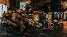 A group of delivery riders on motorbikes is waiting at a traffic light during nighttime in an urban setting. Each rider has a yellow delivery box with the logo 'Glovo' on the back of their bikes. The scene is lit with streetlights, and buildings are visible in the background, including a shop sign for 'Zara'.