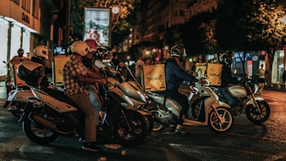 A group of delivery riders on motorbikes is waiting at a traffic light during nighttime in an urban setting. Each rider has a yellow delivery box with the logo 'Glovo' on the back of their bikes. The scene is lit with streetlights, and buildings are visible in the background, including a shop sign for 'Zara'.