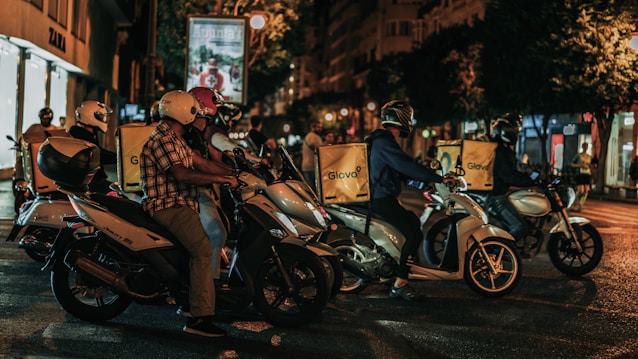 A group of delivery riders on motorbikes is waiting at a traffic light during nighttime in an urban setting. Each rider has a yellow delivery box with the logo 'Glovo' on the back of their bikes. The scene is lit with streetlights, and buildings are visible in the background, including a shop sign for 'Zara'.