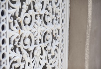 A close-up of an ornate, white wrought iron grill with intricate scrollwork and floral patterns set against a textured, beige wall.