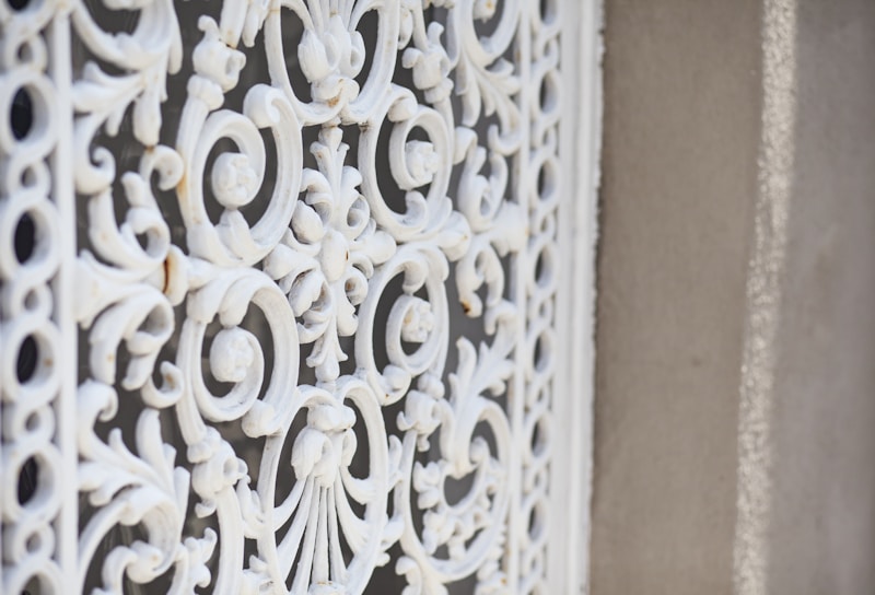 A close-up of an ornate, white wrought iron grill with intricate scrollwork and floral patterns set against a textured, beige wall.