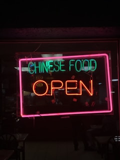 A neon sign is displayed in a window with the words 'Chinese Food' in green and 'Open' in red. The sign is outlined with a bright pink frame, and the background shows part of the interior of a restaurant with dim lighting.