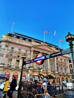 A bustling urban scene featuring a classic London Underground entrance with a bright red and blue sign against a backdrop of a historic building adorned with banners. People are walking by, some carrying cameras, amidst the vibrant signage of various shops and a clear blue sky above.