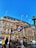 A bustling urban scene featuring a classic London Underground entrance with a bright red and blue sign against a backdrop of a historic building adorned with banners. People are walking by, some carrying cameras, amidst the vibrant signage of various shops and a clear blue sky above.