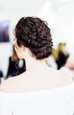 A person with dark, neatly styled hair in an elegant updo, viewed from the back. The hair is intricately curled and pinned, revealing the nape of the neck. The individual is wearing an off-the-shoulder white top, with a softly blurred background suggesting an indoor setting with other people present.