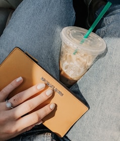 A person wearing blue jeans holds a tan Michael Kors wallet in one hand, with a visible ring on their finger. Next to the wallet, there is an iced coffee in a clear plastic cup with a green straw, displaying the Starbucks logo.