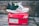 A pair of white and black running shoes with green accents sits on top of a red New Balance shoe box. The shoes feature a distinctive white foam sole and the brand's logo prominently displayed. The background consists of weathered wooden planks, adding a rustic feel to the scene.