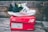 A pair of white and black running shoes with green accents sits on top of a red New Balance shoe box. The shoes feature a distinctive white foam sole and the brand's logo prominently displayed. The background consists of weathered wooden planks, adding a rustic feel to the scene.