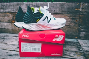A pair of white and black running shoes with green accents sits on top of a red New Balance shoe box. The shoes feature a distinctive white foam sole and the brand's logo prominently displayed. The background consists of weathered wooden planks, adding a rustic feel to the scene.