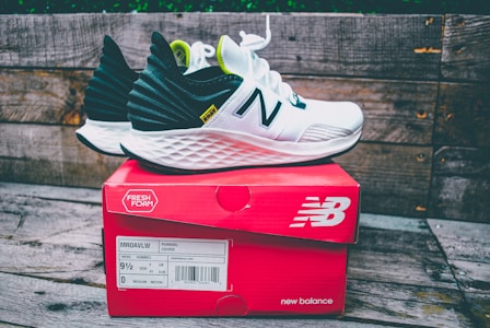 A pair of white and black running shoes with green accents sits on top of a red New Balance shoe box. The shoes feature a distinctive white foam sole and the brand's logo prominently displayed. The background consists of weathered wooden planks, adding a rustic feel to the scene.
