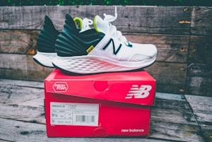 A pair of white and black running shoes with green accents sits on top of a red New Balance shoe box. The shoes feature a distinctive white foam sole and the brand's logo prominently displayed. The background consists of weathered wooden planks, adding a rustic feel to the scene.