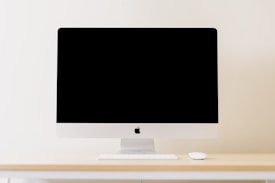 A minimalistic workspace setting featuring a modern computer with a blank screen. The computer is placed on a light wooden desk, accompanied by a wireless keyboard and mouse. The background is a plain, cream-colored wall, enhancing the clean and simplistic aesthetic.