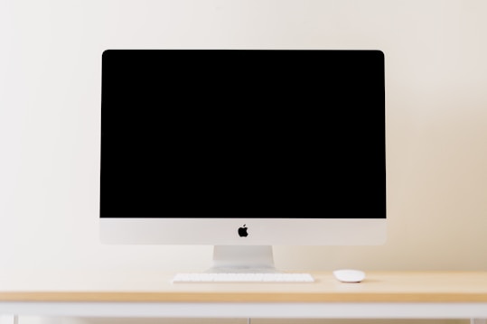 A minimalistic workspace setting featuring a modern computer with a blank screen. The computer is placed on a light wooden desk, accompanied by a wireless keyboard and mouse. The background is a plain, cream-colored wall, enhancing the clean and simplistic aesthetic.
