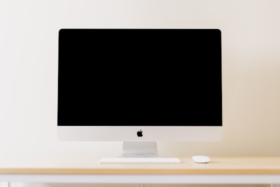 A minimalistic workspace setting featuring a modern computer with a blank screen. The computer is placed on a light wooden desk, accompanied by a wireless keyboard and mouse. The background is a plain, cream-colored wall, enhancing the clean and simplistic aesthetic.