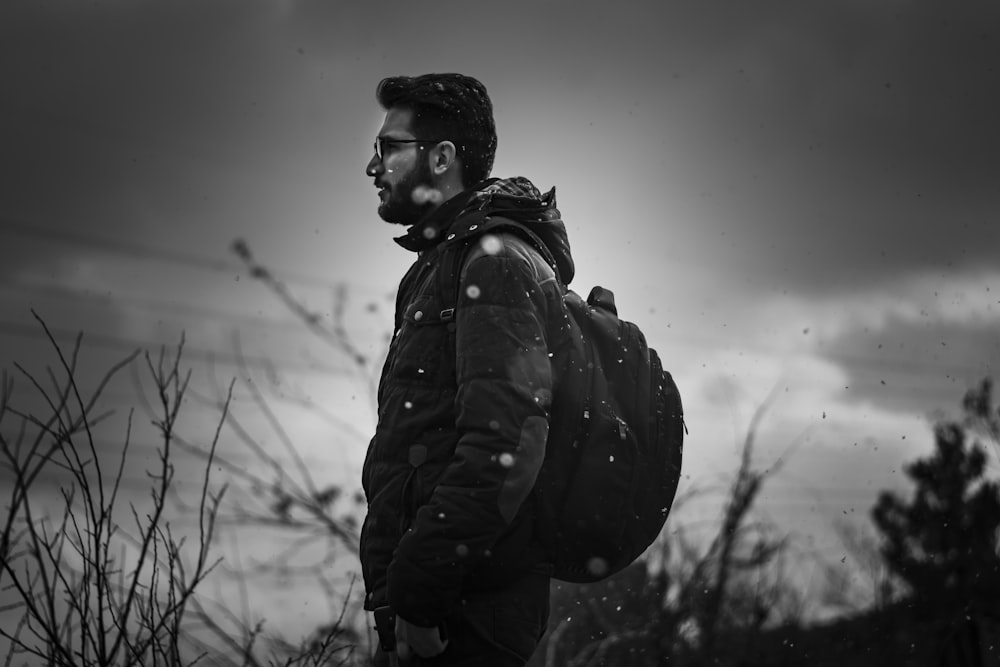 grayscale photography of standing man carrying backpack and wearing sunglasses