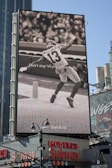 A large billboard displays a black and white image of a football player with the number 13 and the name visible on the jersey. Text on the billboard reads 'Don't stop 'til you get enough' and 'Just do it.' This advertisement occupies a prominent position above a busy urban street corner with stores and signs below.