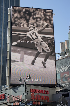 A large billboard displays a black and white image of a football player with the number 13 and the name visible on the jersey. Text on the billboard reads 'Don't stop 'til you get enough' and 'Just do it.' This advertisement occupies a prominent position above a busy urban street corner with stores and signs below.
