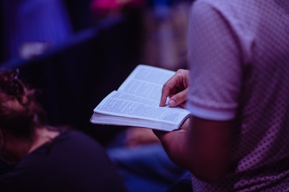 A person is seen holding and reading an open book, focusing on the text. The environment is dimly lit, suggesting an intimate or quiet setting. The person's attire includes a patterned short-sleeved shirt.