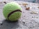 A worn-out tennis ball with visible scuff marks rests on a rough, sandy surface, surrounded by dry leaves and small debris. The background is blurred, emphasizing the texture of the ball and the ground.