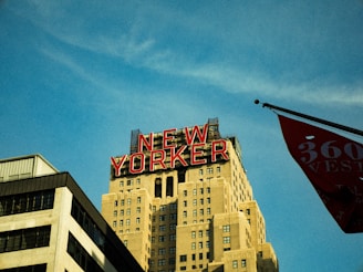 A skyline view featuring a tall building with a classic Art Deco style and a prominent red neon sign displaying the words 'NEW YORKER' at the top. The structure is surrounded by a clear blue sky and a partial view of another building and a flag in the foreground.