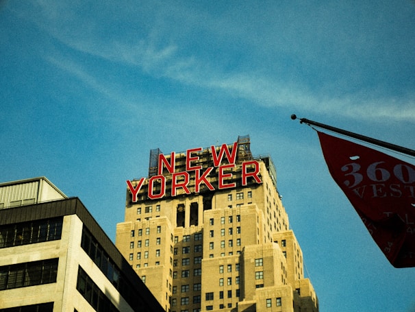 A skyline view featuring a tall building with a classic Art Deco style and a prominent red neon sign displaying the words 'NEW YORKER' at the top. The structure is surrounded by a clear blue sky and a partial view of another building and a flag in the foreground.