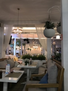 A cozy and elegantly designed restaurant interior features soft lighting, wooden furniture, and hanging plants. The space includes white sheer curtains, decorative light fixtures, and a variety of cushions on the seating. Tables are set with placemats and small arrangements of flowers.