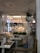 A cozy and elegantly designed restaurant interior features soft lighting, wooden furniture, and hanging plants. The space includes white sheer curtains, decorative light fixtures, and a variety of cushions on the seating. Tables are set with placemats and small arrangements of flowers.