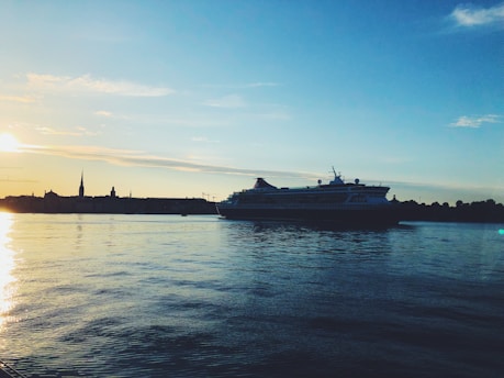 A large cruise ship sails on calm water with a silhouette of a city skyline in the background. The setting sun gives the scene a warm glow, with the sky displaying hues of blue and orange. The water reflects the colors of the sky, adding to the serene atmosphere.