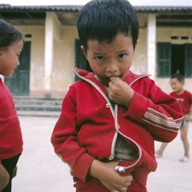 A young child stands in the foreground, wearing a red jacket. The expression is somewhat serious, with the child biting their fingernail. In the background, there are other children, and an old, weathered building with green doors and windows.