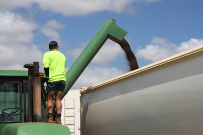 A person stands on a green tractor, wearing a bright yellow-green high-visibility shirt and shorts. The tractor is parked next to a large container where grain is being poured through a green chute. The sky is partly cloudy.