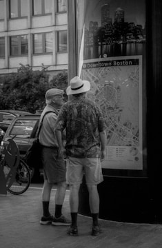 Two people, both casually dressed and wearing hats, are standing in front of a large map titled Downtown Boston. They seem to be examining the details of the map, with buildings and trees visible in the background. The scene is in black and white, giving it a classic or nostalgic feel.