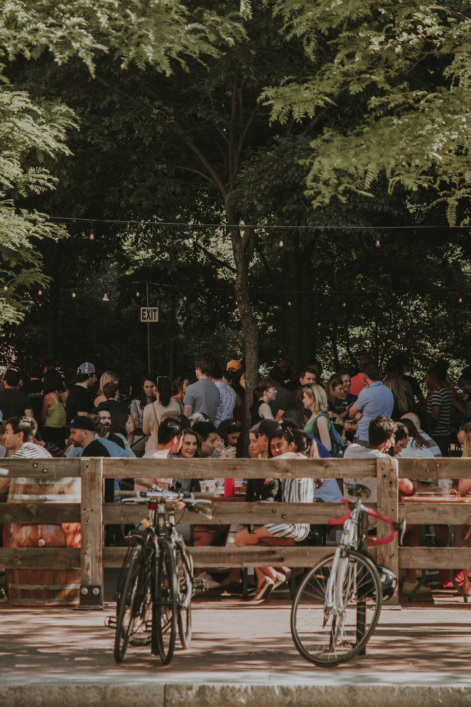 The image depicts a lively scene at an outdoor event, with numerous people gathered around and engaged in various activities. The focal point is two bicycles parked on the sidewalk next to a wooden fence, drawing attention from passersby who are drawn to their unique design. In the background, there's a crowd of people standing near trees, adding to the festive atmosphere.
