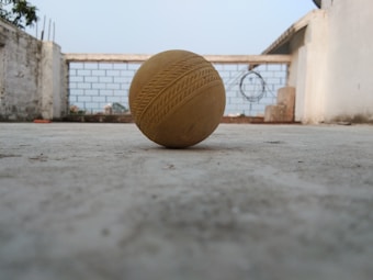 A yellow cricket ball with seam patterns rests on a concrete surface in an open area. In the background, there is a low, brick wall with wire fencing, and parts of a structure are visible. The scene is outdoors, suggesting a rooftop or a courtyard.