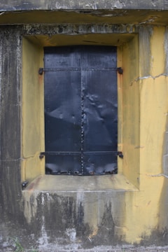 A black metal door or window cover is set within a rough yellow concrete wall, showing signs of rust and wear. The surface surrounding the window is stained and cracked, adding to a sense of age and neglect.
