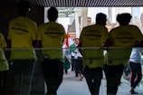 Several people gather in a hallway, where some are wearing bright yellow shirts labeled 'VOLUNTEERS'. Others in the background carry the Indian national flag, creating a sense of cultural celebration.