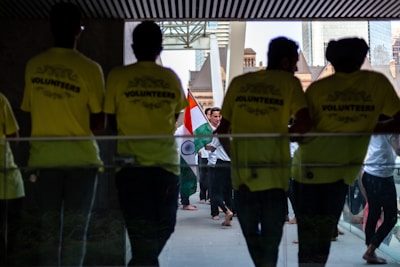 Several people gather in a hallway, where some are wearing bright yellow shirts labeled 'VOLUNTEERS'. Others in the background carry the Indian national flag, creating a sense of cultural celebration.