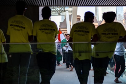Several people gather in a hallway, where some are wearing bright yellow shirts labeled 'VOLUNTEERS'. Others in the background carry the Indian national flag, creating a sense of cultural celebration.