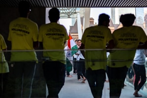 Several people gather in a hallway, where some are wearing bright yellow shirts labeled 'VOLUNTEERS'. Others in the background carry the Indian national flag, creating a sense of cultural celebration.