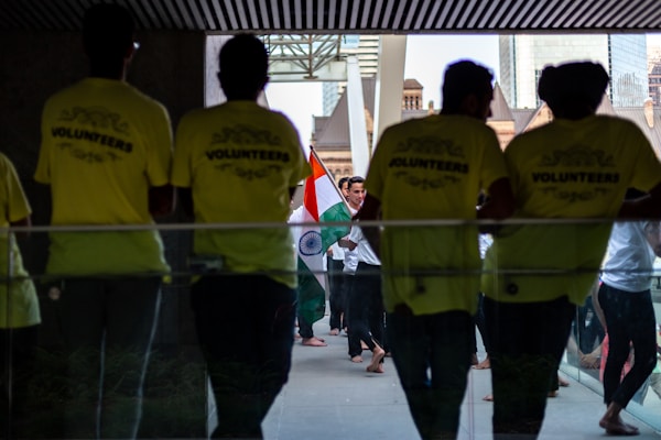 Several people gather in a hallway, where some are wearing bright yellow shirts labeled 'VOLUNTEERS'. Others in the background carry the Indian national flag, creating a sense of cultural celebration.