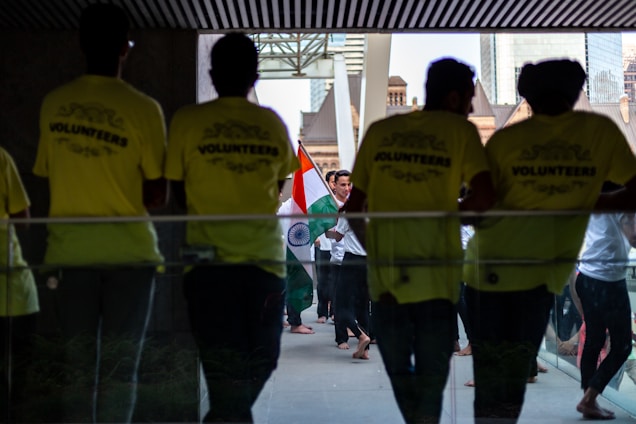 Several people gather in a hallway, where some are wearing bright yellow shirts labeled 'VOLUNTEERS'. Others in the background carry the Indian national flag, creating a sense of cultural celebration.