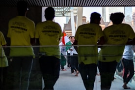Several people gather in a hallway, where some are wearing bright yellow shirts labeled 'VOLUNTEERS'. Others in the background carry the Indian national flag, creating a sense of cultural celebration.